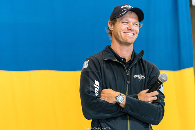 A man dressed in a black top and baseball club stands in front of a yellow and blue background